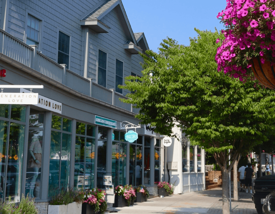 A street view of shops with large windows and vibrant flower pots under clear blue skies.