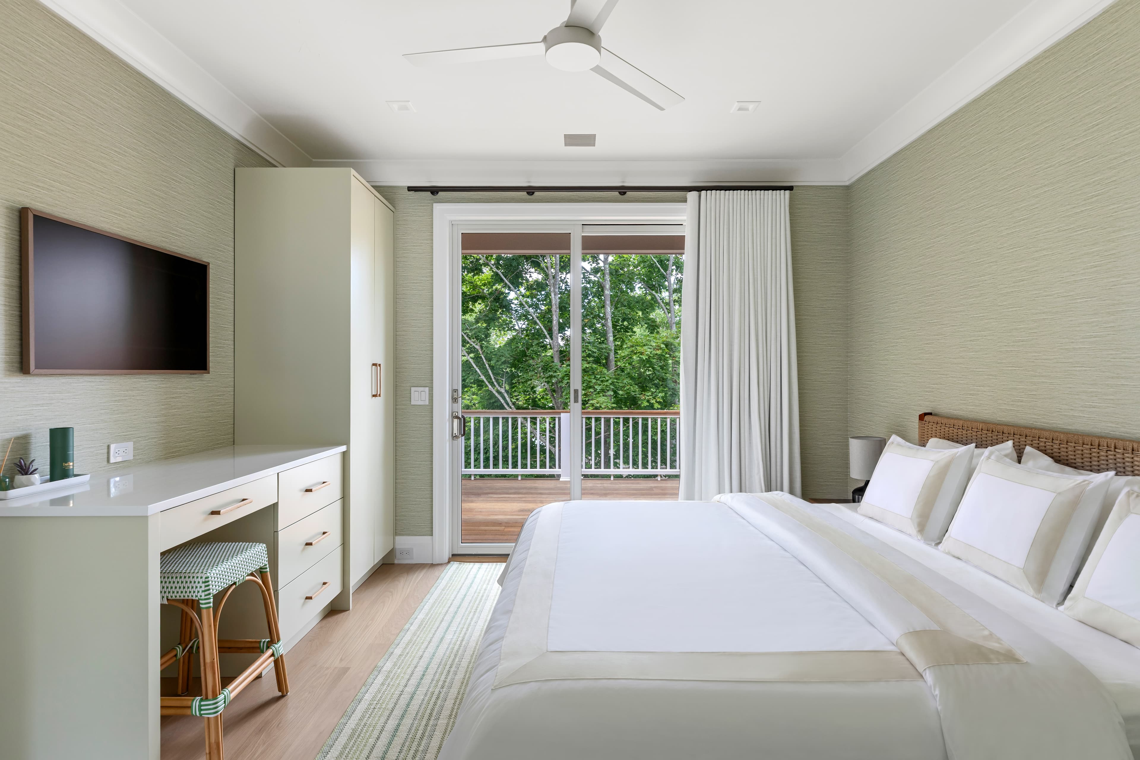 A modern bedroom featuring a large bed, light green walls, a dresser, and a view of a balcony with trees outside.