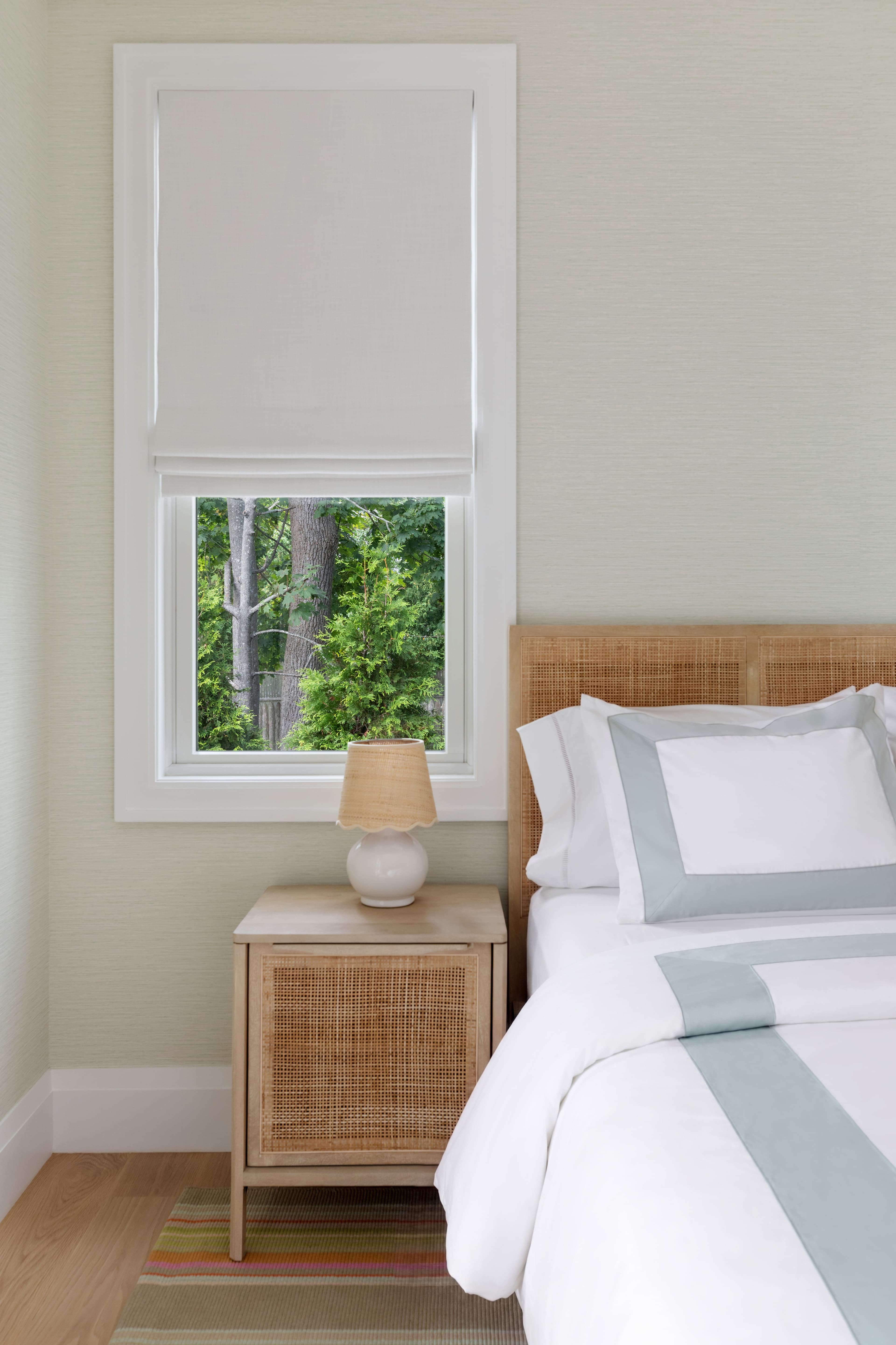 A cozy bedroom with a bed, nightstand, lamp, and a window displaying greenery outside.