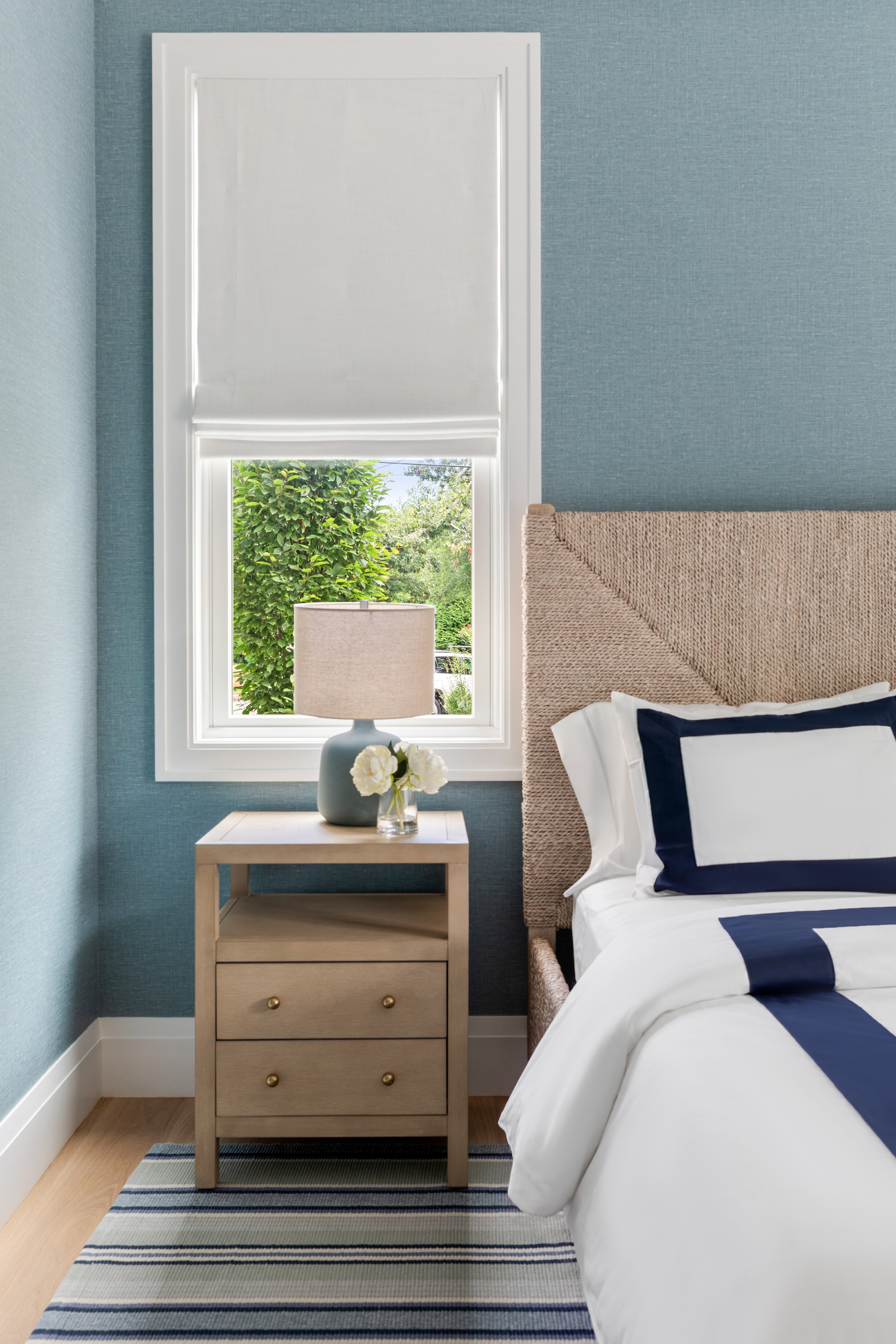 A modern bedroom featuring a textured headboard, a bedside table with a lamp and vase, and a window with light blue walls.