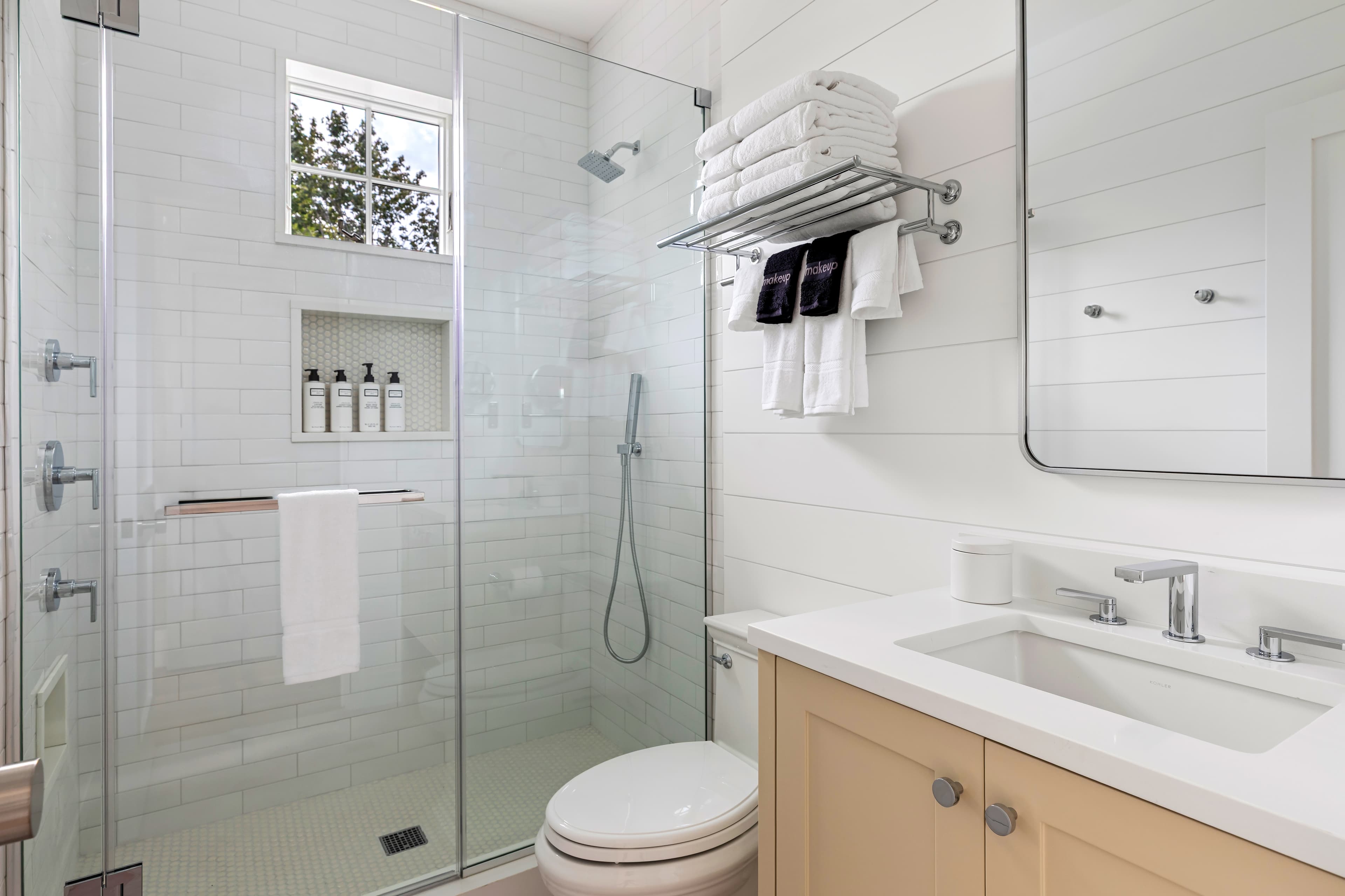 Modern bathroom featuring a glass shower, white tiles, a mirrored sink area, and neatly arranged towels.