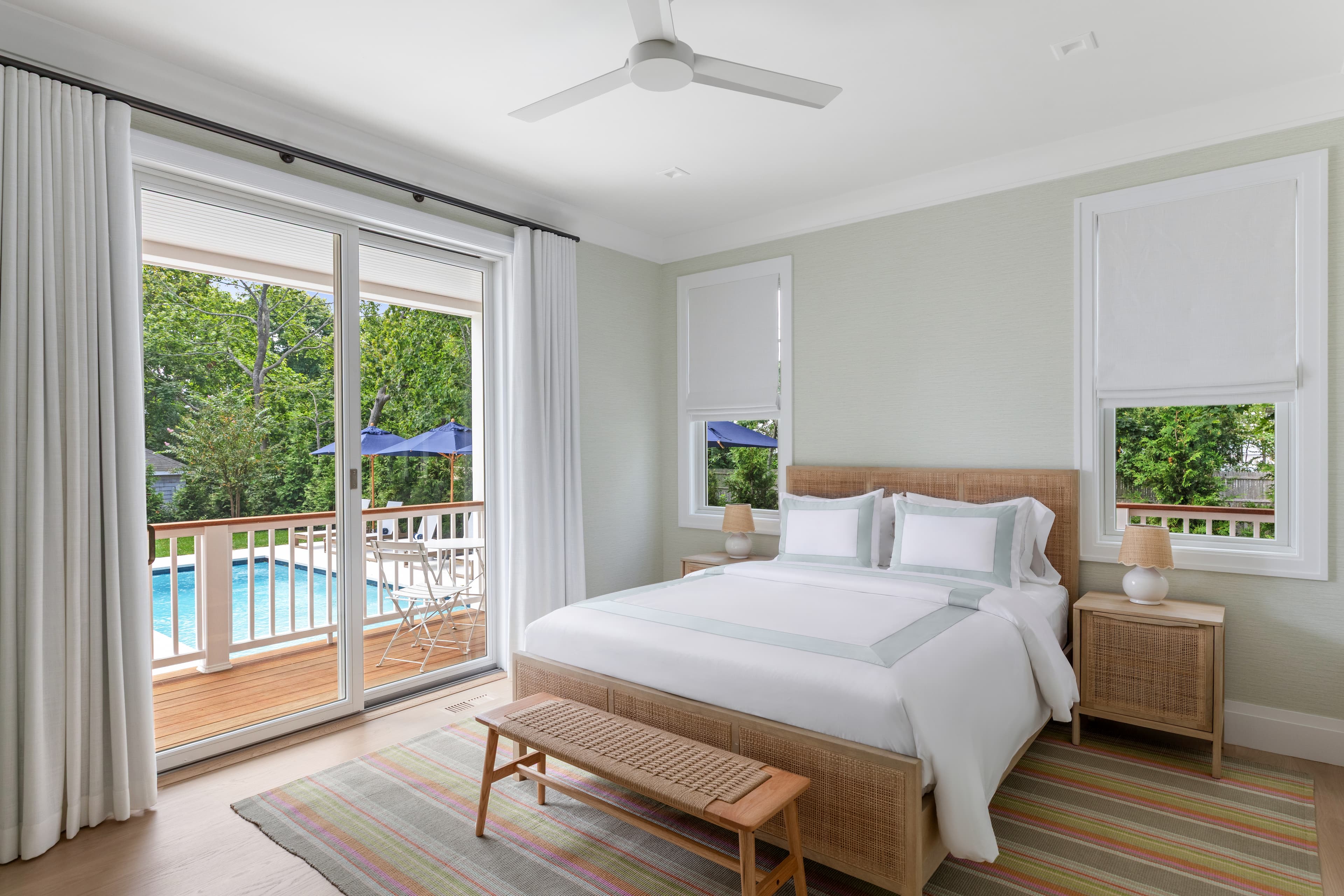 A bright, modern bedroom featuring a large bed, a wooden bench, and windows overlooking a pool area.
