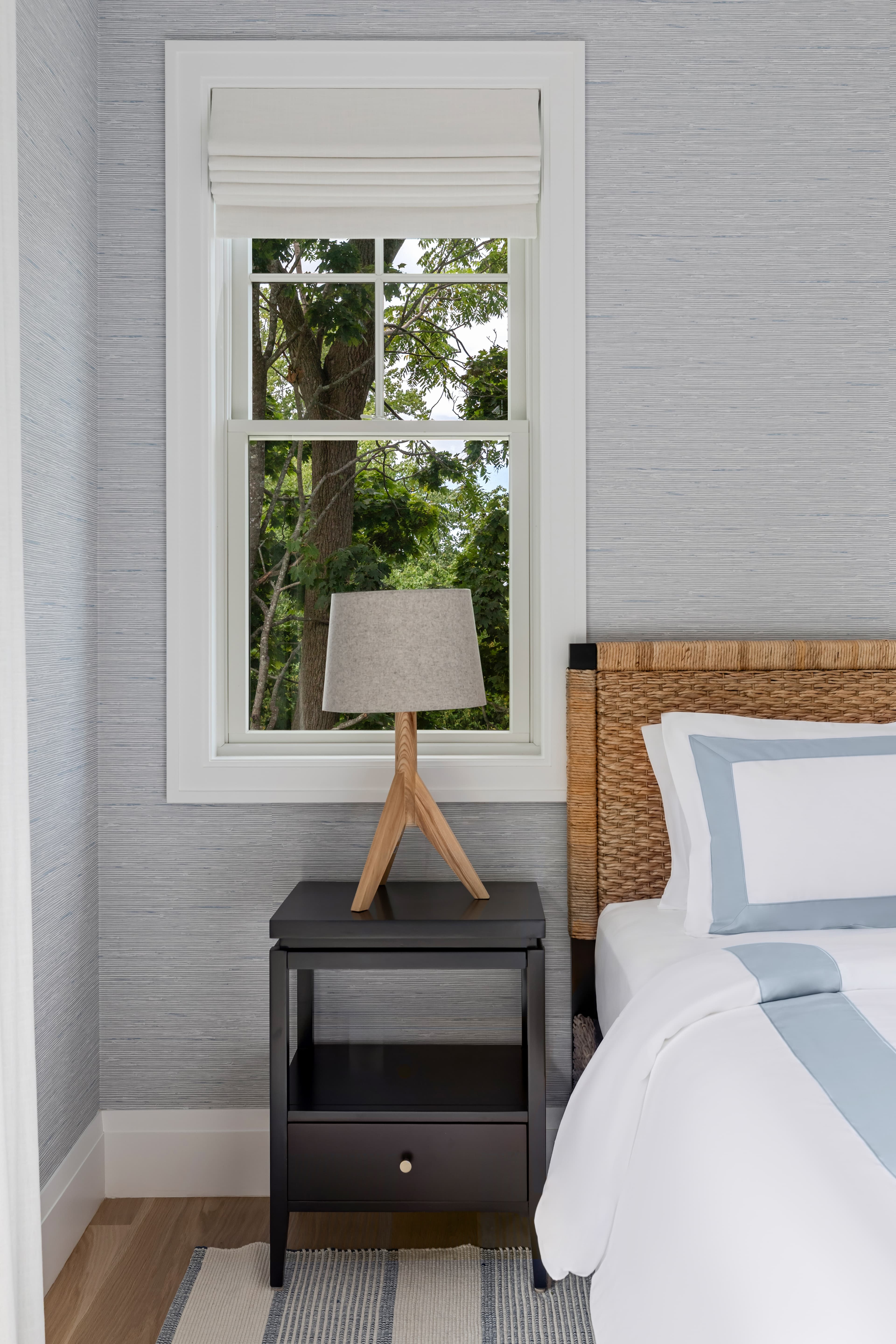 A cozy bedroom scene featuring a bedside table with a lamp beside a bed and a window showcasing greenery outside.