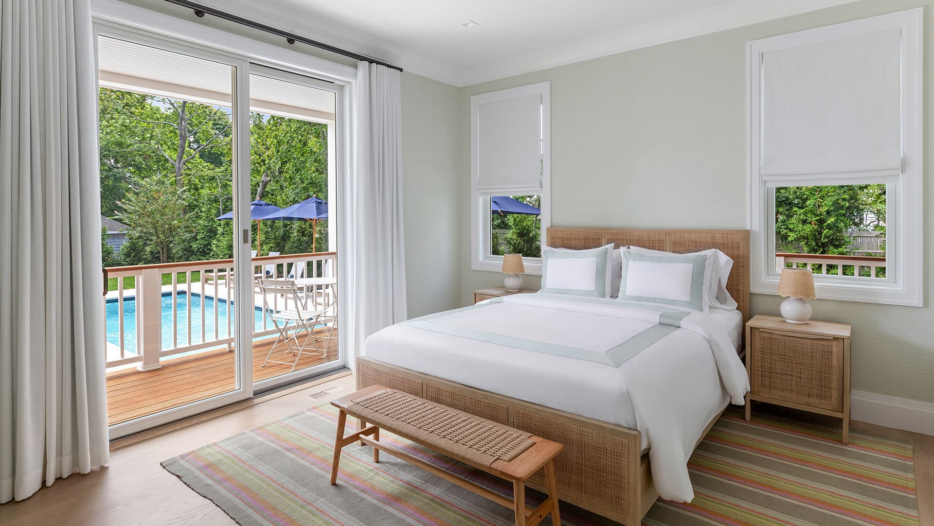 A bright, modern bedroom with a large bed, a bench, and sliding doors opening to a deck by a pool.