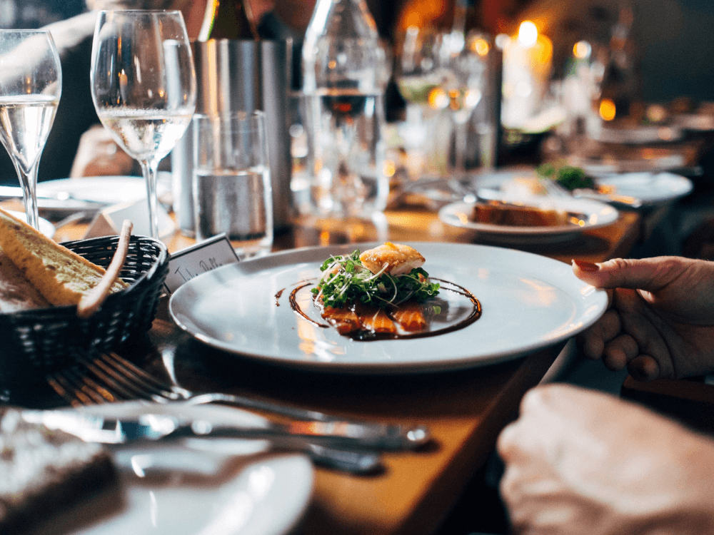 A beautifully plated dish garnished with greens is being served at a dining table set with wine glasses and candlelight.