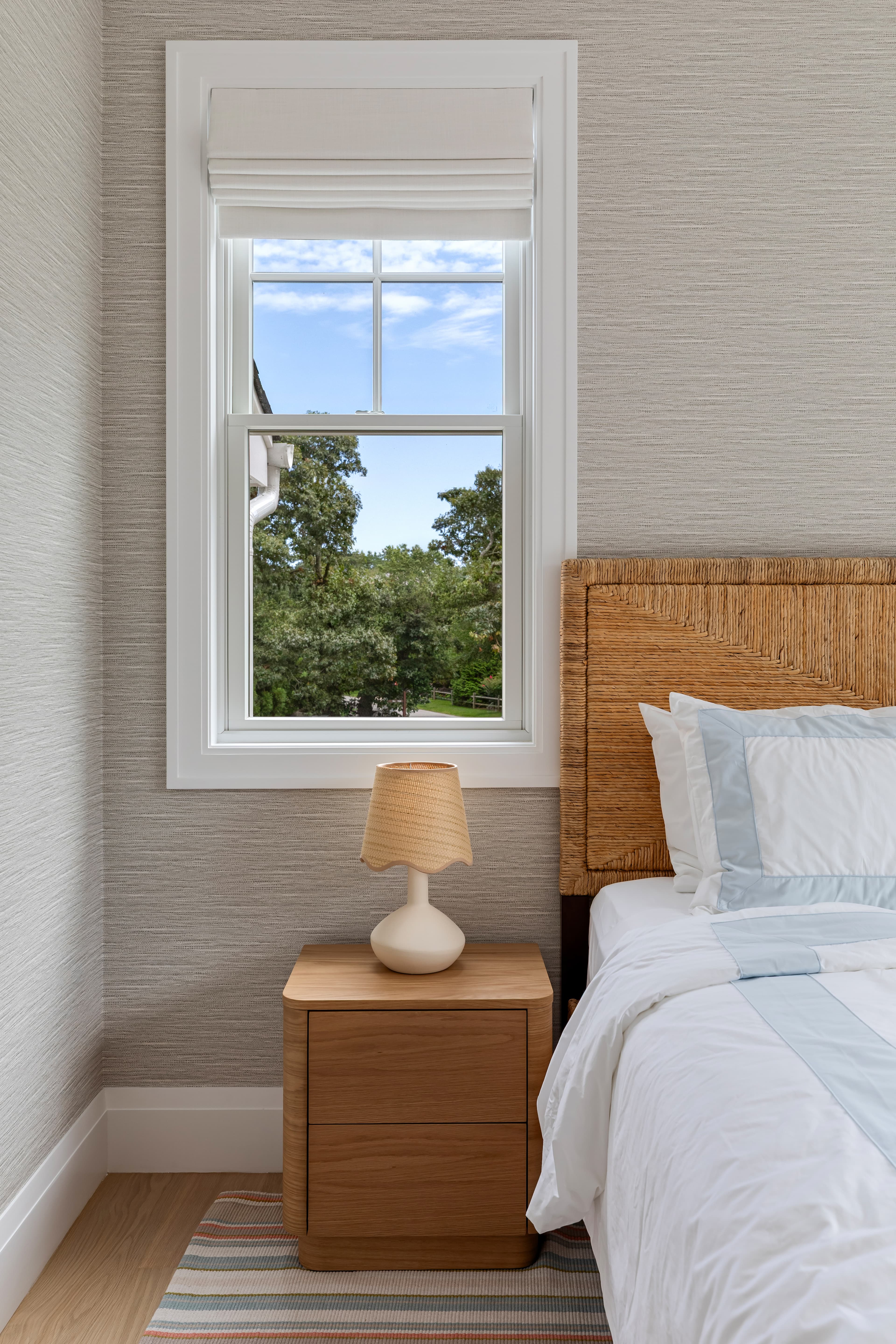 A cozy bedroom featuring a woven headboard, bedside table with a lamp, and a window view of greenery.