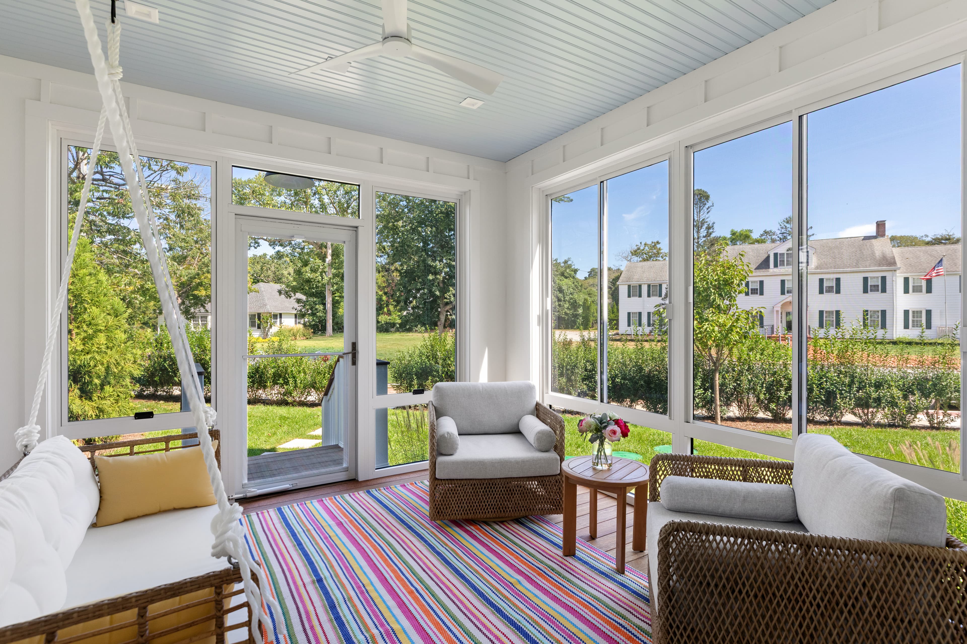 Bright sunroom with large windows, two armchairs, a white couch, and a colorful striped rug.