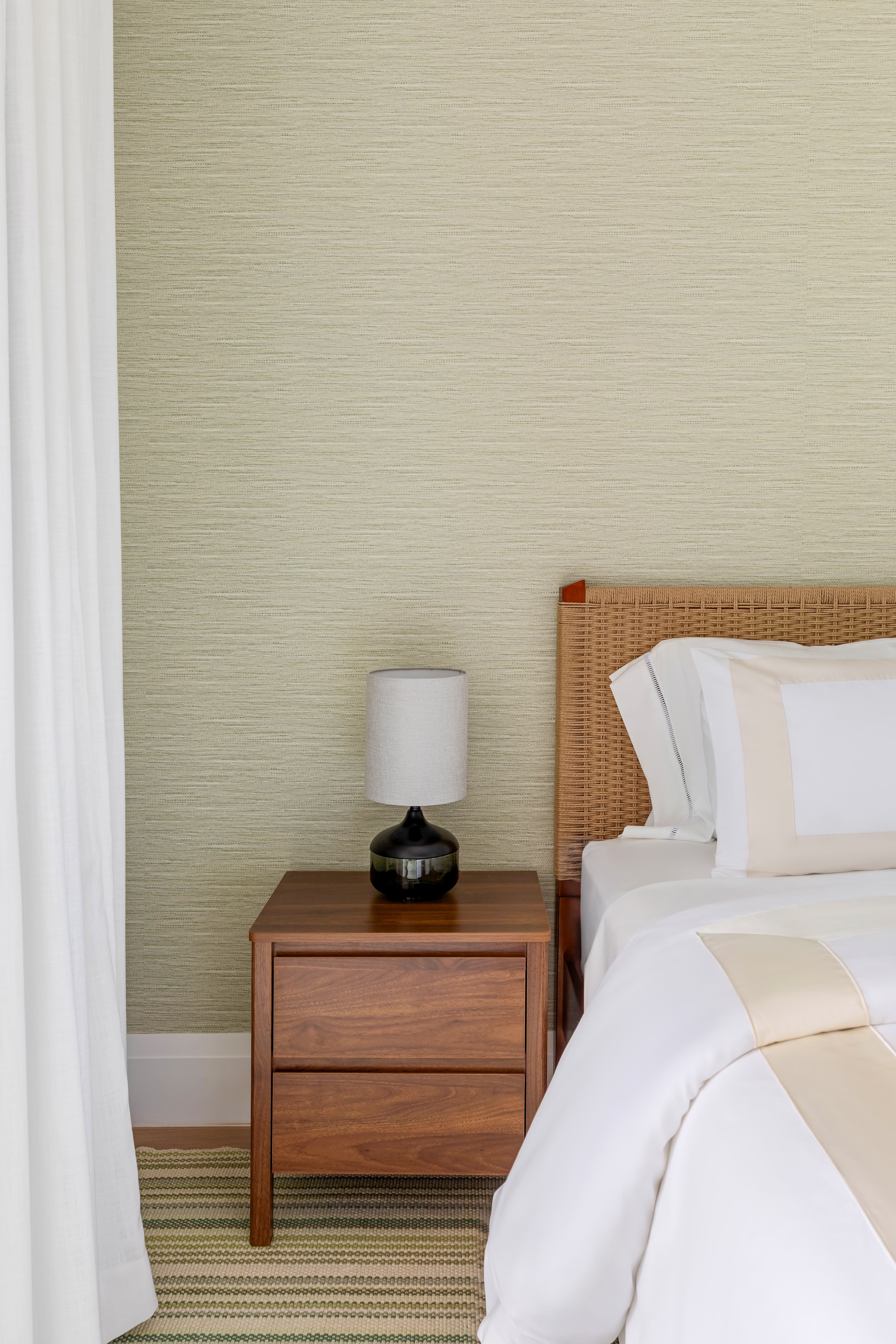 A cozy bedroom corner featuring a wicker headboard, white bedding, a wooden nightstand with a black lamp, and a textured wall.