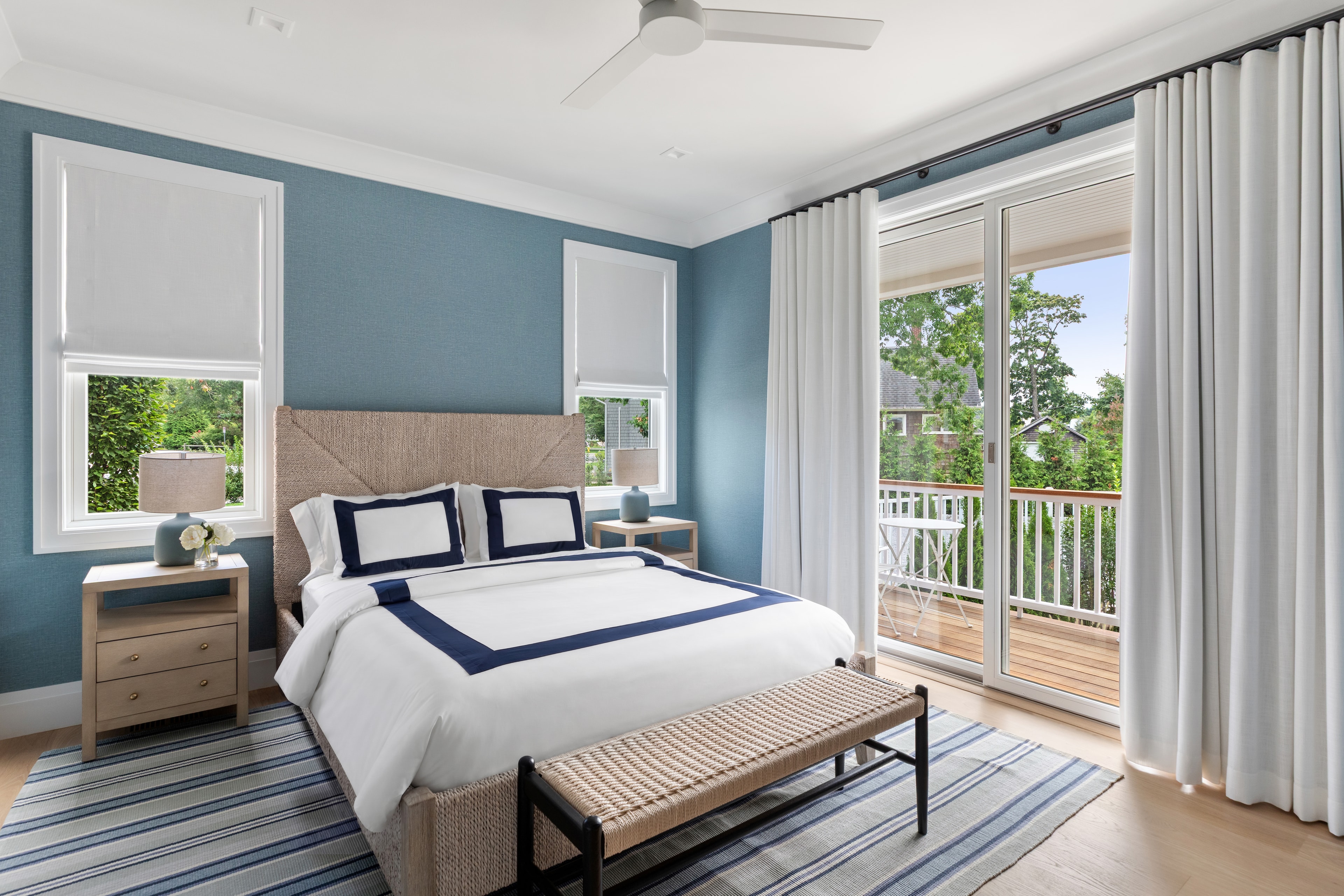 A modern bedroom with a blue accent wall, a queen-sized bed, bedside tables, and large windows overlooking a balcony.